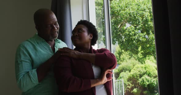 African american senior couple embracing each other while looking out of the window at home