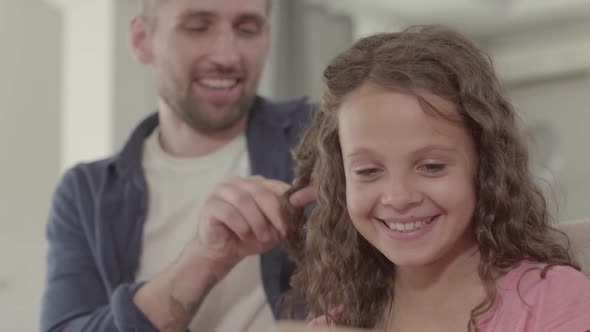 The Father Braids the Girl's Pigtail, Smiling. The Man Spending Time with His Cute Little Daughter