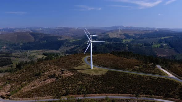 Spinning wind turbines lined up on top of a hill with forests connected by roads and many more wind