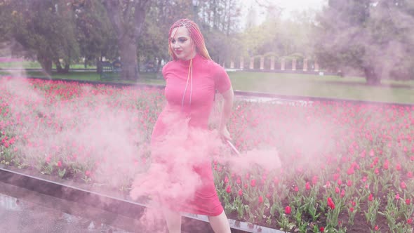 A Girl with Makeup with Rainbow Braids in Red Dress Posing in Red Smoke Against the Background of a