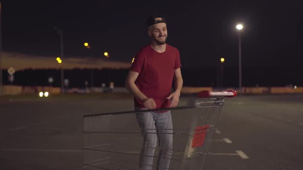 Laughing Cheerful Caucasian Man Having Fun with Shoppng Cart on Empty Parking Lot at Night. Portrait