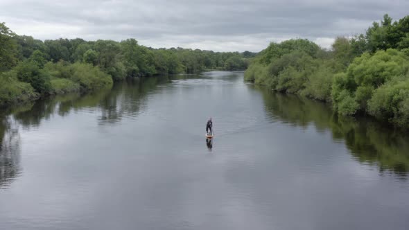 Standup paddleboarder paddles toward camera on green country river