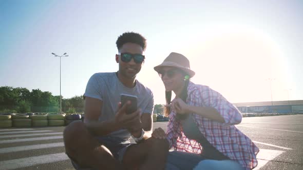 Young Mixed Race Hipster Couple Listening Music Outdoors While Sitting on Longboards