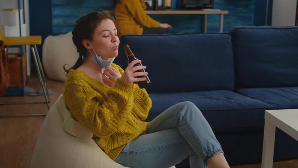 Relaxed Woman Taking Off Face Mask Looking at Camera Drinking Beer