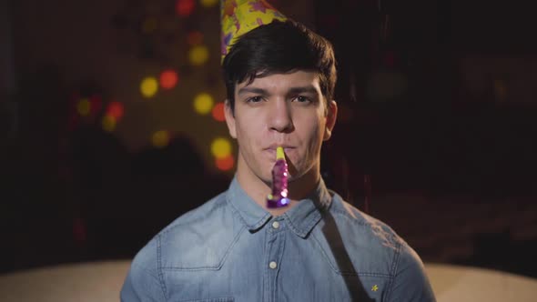 Portrait of Young Man in Birthday Hat Looking in the Camera Blowing Noisemaker