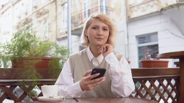 Young Blonde Woman with Elegant Manicure in a Beige Vest and White Blouse Holding a Cell Phone While