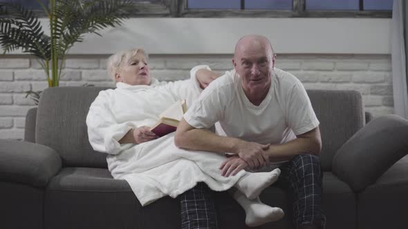 Mature Caucasian Man Sitting on the Couch with Wife and Watching Sports Match on TV. Woman Reading