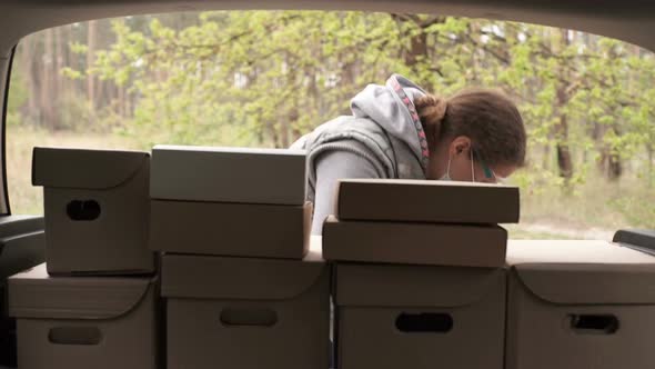 A Volunteer in a Protective Suit Loads Boxes Into the Trunk of a Car