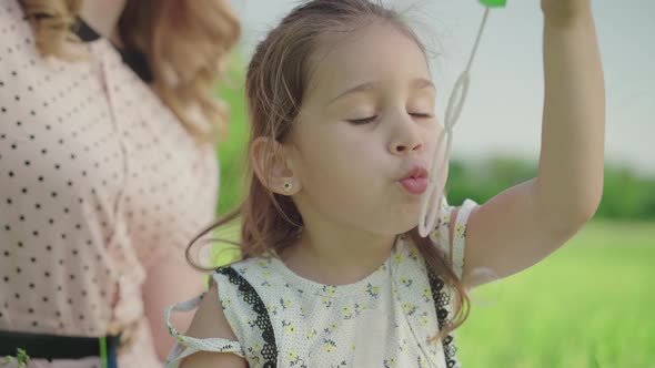 Close-up of Positive Little Girl Blowing Soap Bubbles As Unrecognizable Mother Clapping Hands at the