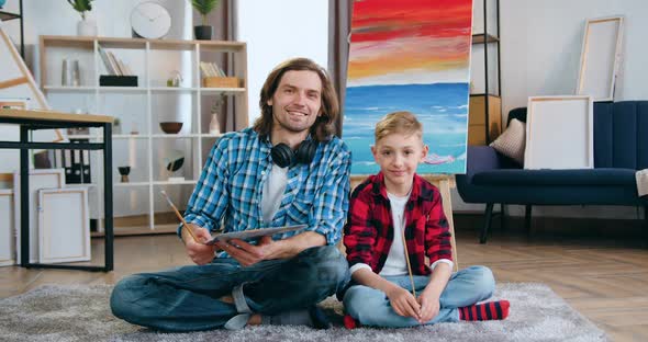 Man and Cute Boy Both Artists Sitting in front of Camera Near Painted Picture on canvas