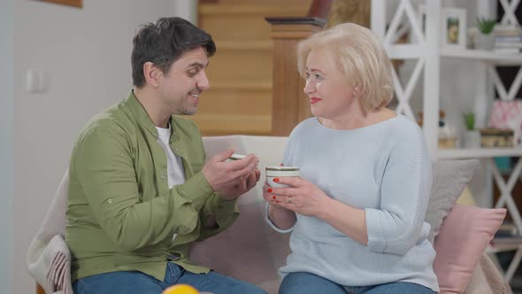 Positive Adult Son Talking with Senior Mother Holding Herbal Tea Cups