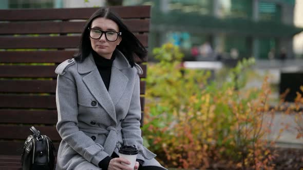 a Brunette in a Gray Coat Glasses with a Bag and a Paper Cup in Hands is Sitting on a Wooden Bench