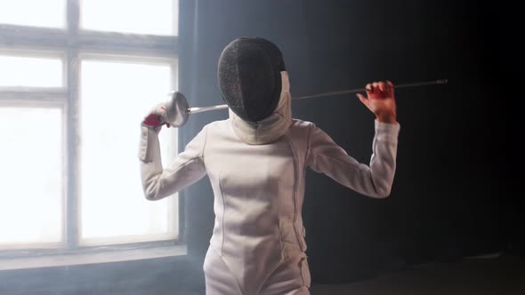 A Young Woman Fencer with a Helmet Putting the Sword Behind Her Shoulders in the Studio