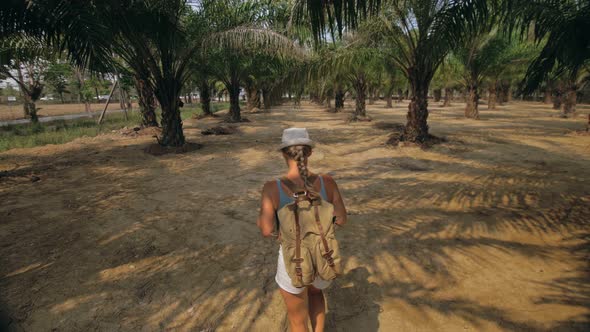 Woman Tourist with Plait Walks Looking Around at Growing Young Trees with Lush Leaves at Oil Palm