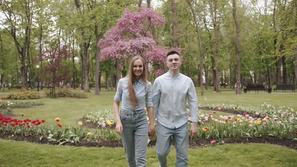 Young Romantic Couple Hold Hands and Goes Against the Background of Flowers in the Park