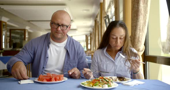 Husband Bald and Glasses and Dark-haired Middle-aged Wife, Sitting in a Cafe, Eat, Communicate. In