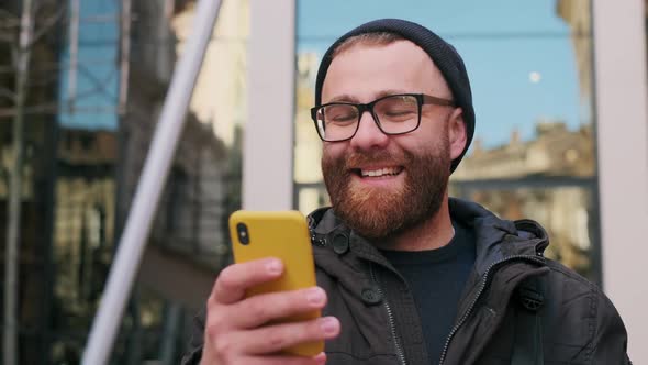 Close Up View of Bearded Man in 30s Looking at Phone Screen and Laughing While Walking at Street