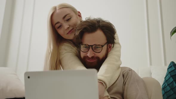 Affectionate Caucasian Couple Sitting in the Living Room and Browsing the Internet Using a Silver