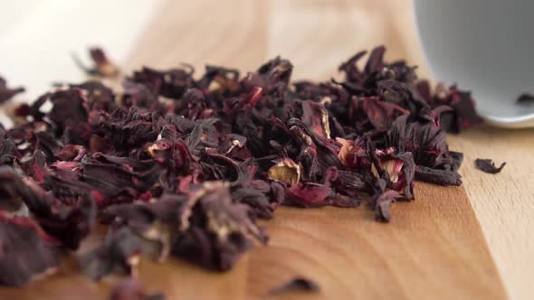Red dry hibiscus tea falls out of a white cup on a wooden surface. Pink dried flower petals