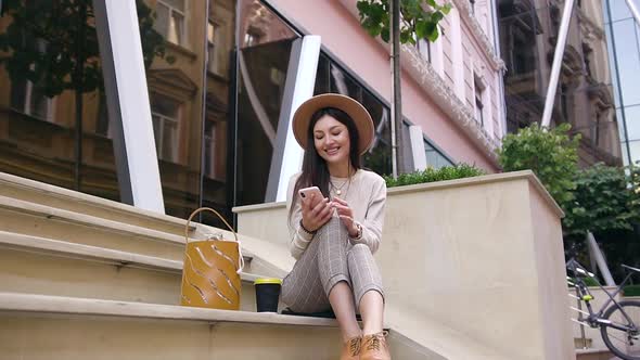 Smiling Young Lady in Modern Clothes and Trendy Hat Sitting on Steps Near Urban Buildings