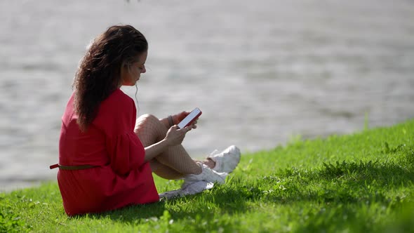 Brunette Woman is Relaxing on River Shore and Using Smartphone Surfing Internet at Nature