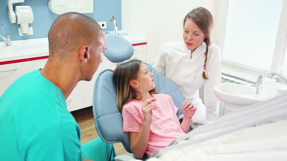 Little girl sitting in the dental chair, talking with dentist and assistant