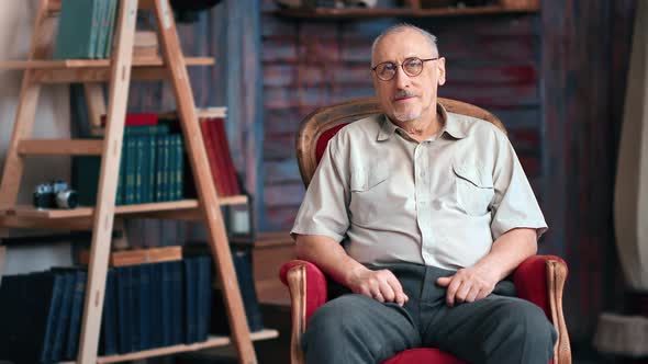 Elderly Smiling Man Sitting on Vintage Armchair at Rustic Home Interior Wooden Bookshelf Library