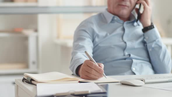 Hand of Elderly Businessman Writing in Journal and Talking on Mobile Phone