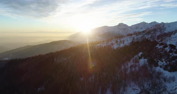 Backward Aerial Top View Over Winter Snowy Mountain and Woods Forest at Sunset or Sunrise