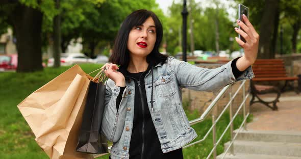 Brunette woman holding paper shopping bags and taking selfie by smartphone in park