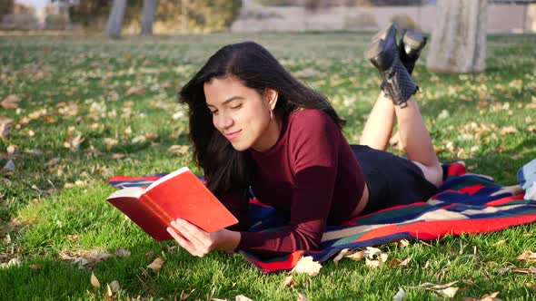 A young woman reading a book in the park during the autumn season with fall leaves blowing in the wi