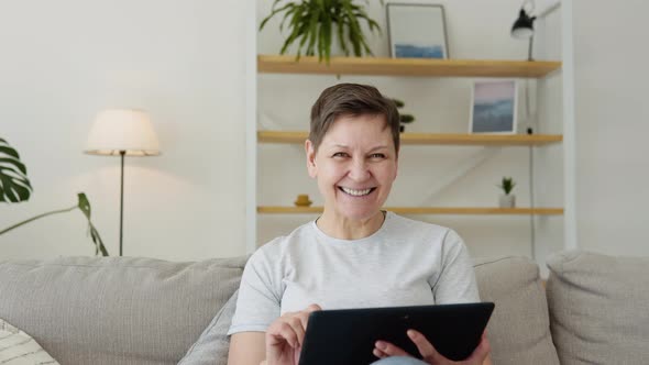 Smiling Elderly Mature Woman Resting on Sofa Using Digital Tablet Alone at Home