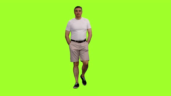 A Man in White T-shirt Walks with Hands in Pockets on Green Background