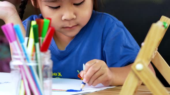 A cute little girl is drawing a rainbow on paper with colored pencils at the table at home.