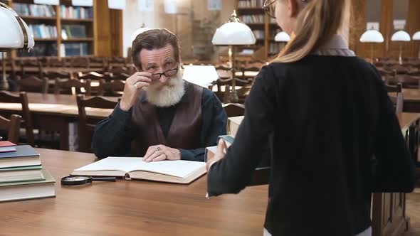 Senior Bearded Man in Glasses Sitting at the Library Table While His Nice Teen Granddaughter