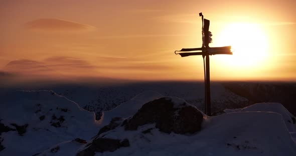 Iron wrought christian cross on top of a mountain over winter. Sun in the orange sky, sunset