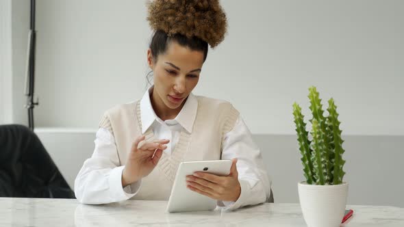 Portrait of a Cheerful Stylish Black Woman in Casual Clothes Sitting at a Desk Using a Tablet in the