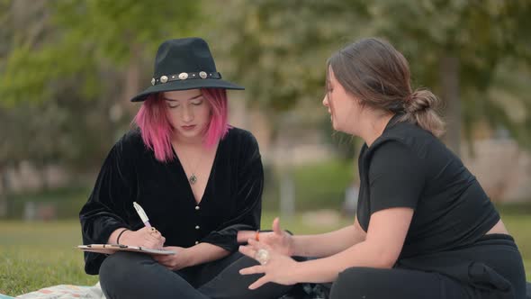 Pink Haired Girl Takes Notes in Notepad While Her Friend Dictates to Her