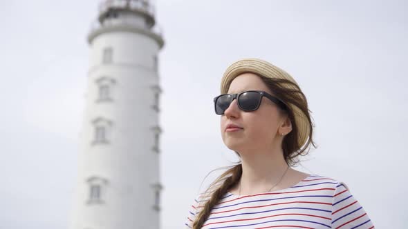 Portrait of a Young Woman in Sunglasses and a Straw Hat Against the Backdrop of the Lighthouse