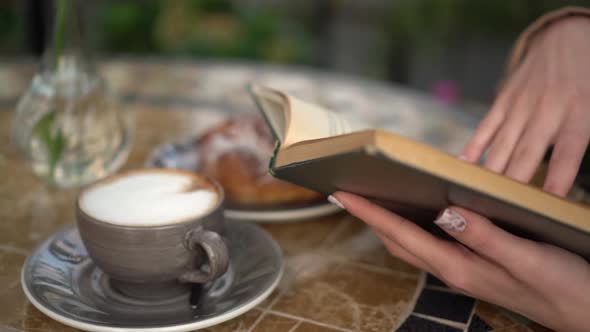 Young Business Woman Sitting in a Cafe and Reading a Book Close-up. Girl with Coffee and a Bun on