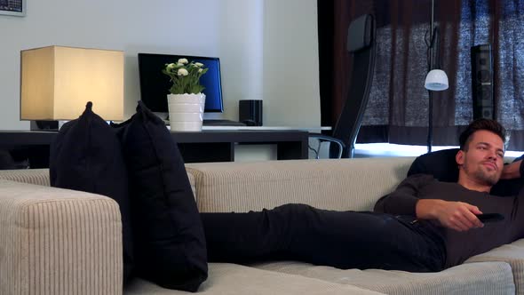 A Young, Handsome Man Lies on a Couch in a Cozy Living Room Watches a TV Which Is Off the Camera