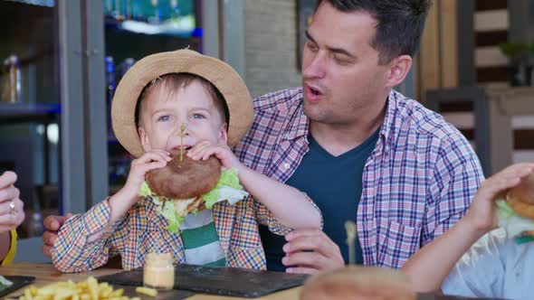 Fast Food, Small Attractive Boy with Straw Hat Eats Mouth Watering Juicy Burger Sitting By Joyful