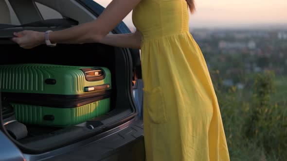Young Woman in Yellow Summer Dress Taking Green Suitcase From Car Trunk
