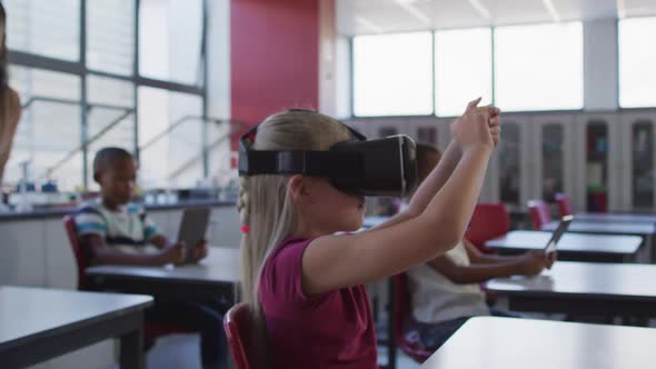 Caucasian schoolgirl sitting at desk in classroom wearing virtual reality glasses and gesticulating