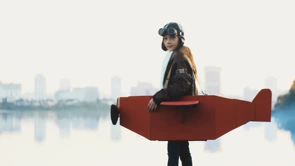 Little Aviator Girl at Big City Lake in Cardboard Plane Playing Pilot, Looking Back and Wearing Old