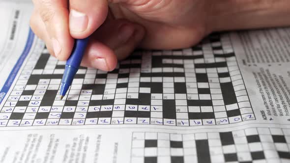 Man solves a crossword clue and writes it down with a blue pen. Crossword is from a local english ne