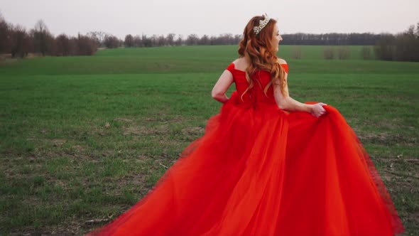 Young Woman in a Luxurious Red Dress Runs Along the Green Field and Smiles