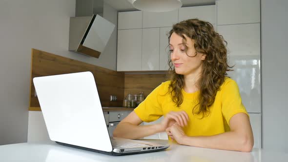 Young Woman with Curly Hair and Yellow Shirt is Working From Home Using Her Laptop at the Kitchen