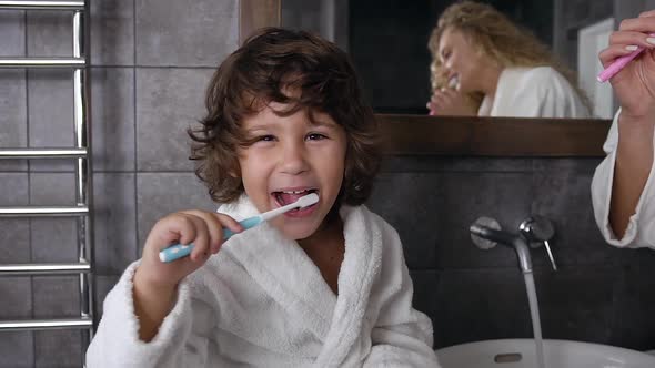 Cheerful Little Boy with Curly Hair in a White Coat is Brushing His Teeth with Toothpaste
