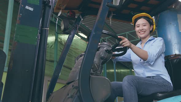 Happy smiling Asian female woman worker in safety helmet driving forklift in warehouse factory.